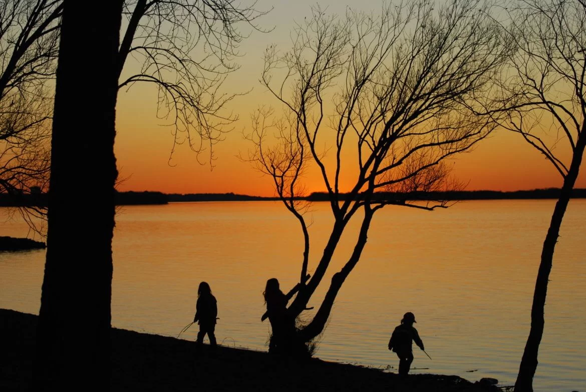 three people walking along the shore of a lake at sunset