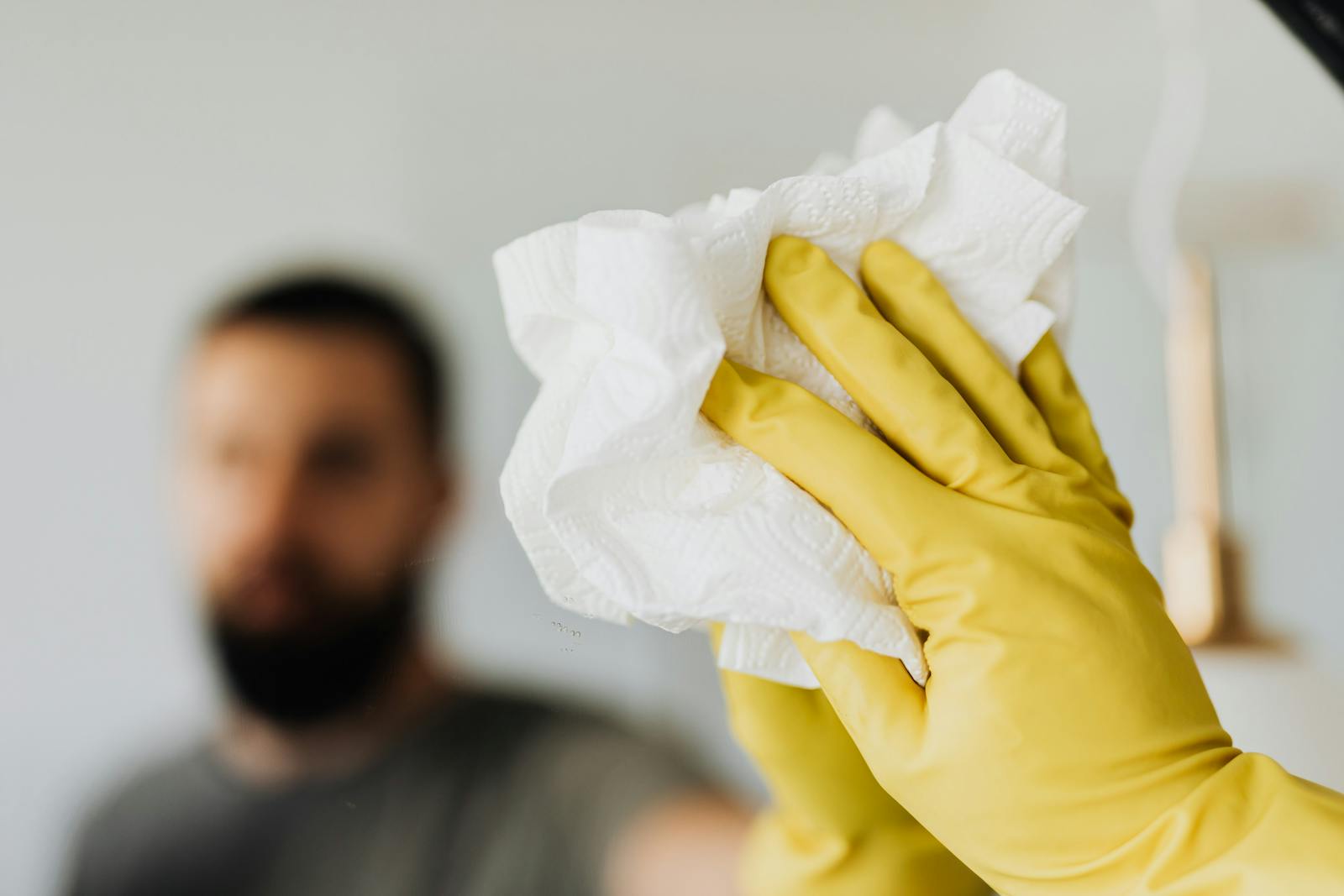 Unrecognizable bearded male wearing yellow latex gloves wiping mirror with napkin in bathroom at home